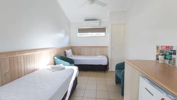 A small, clean room with two single beds, a wooden headboard, a ceiling fan, a small kitchenette, and a window with blinds.
