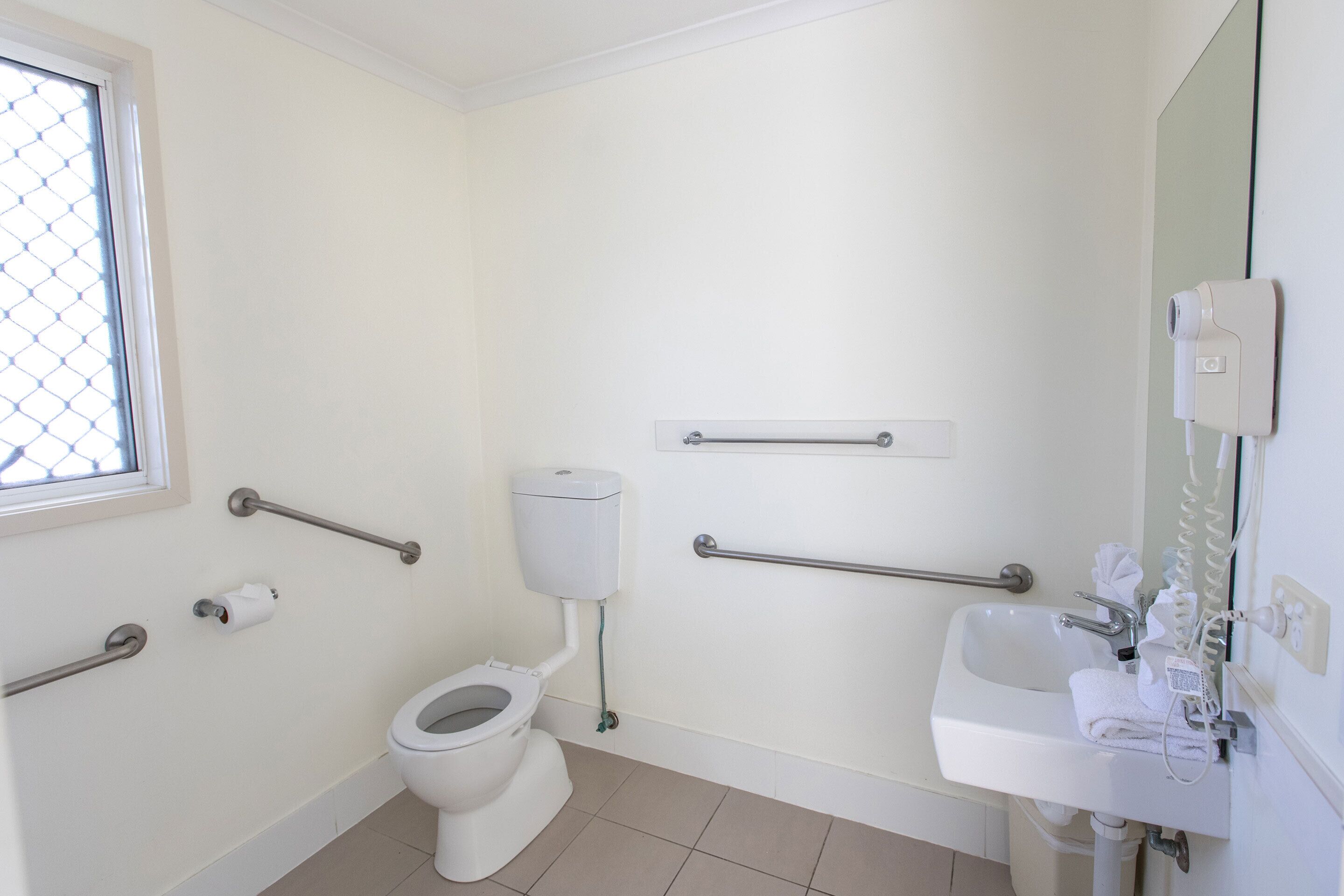 A bathroom with a toilet, handrails, a sink, and a window.