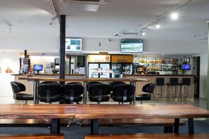 Una zona de bar con una larga mesa de madera y varias sillas, un letrero de área de servicio y una pantalla de televisión mostrando un evento deportivo.