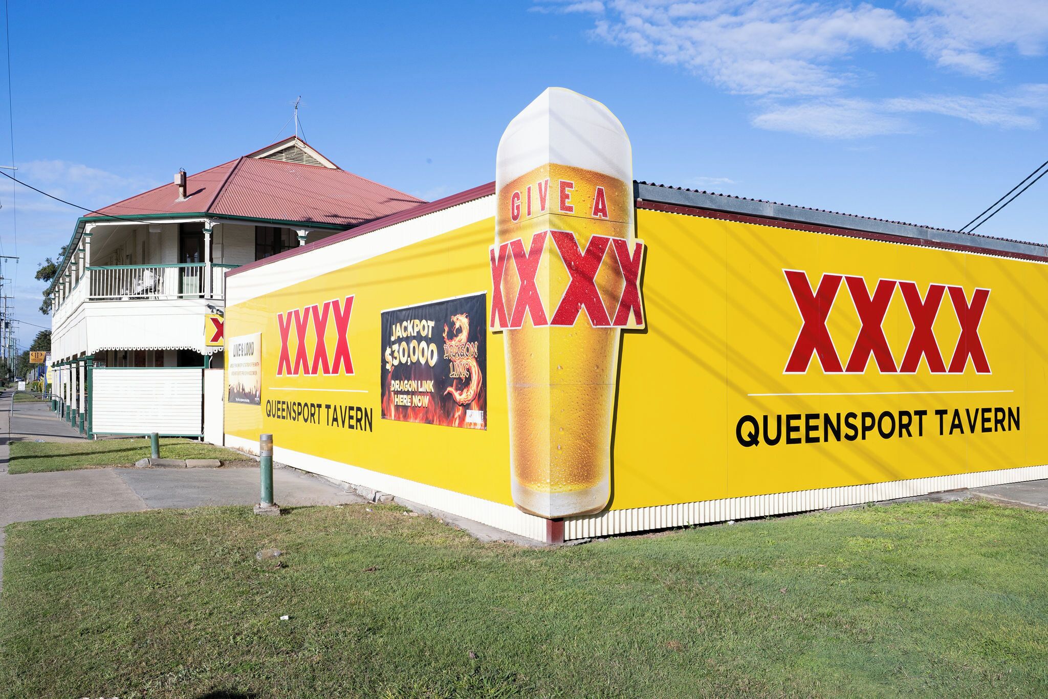 Ein Gebäude mit einer auffälligen gelben Wand, die große, rote 'XXXX'-Schriftzüge und eine Bierglas-Grafik zeigt. Auf dem Gebäude steht 'Queensport Tavern'.