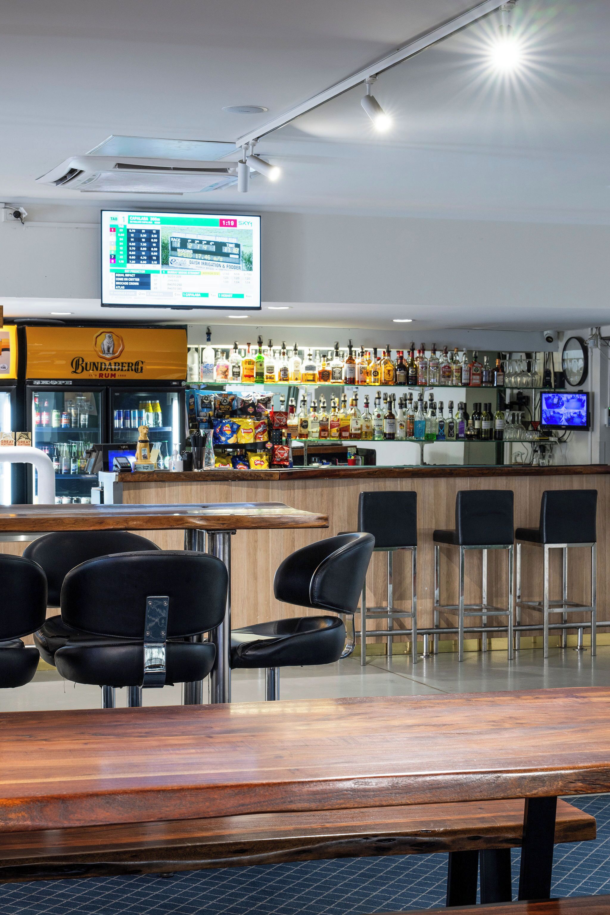 Un espace bar doté d’un écran numérique affichant les résultats sportifs, d’une variété de bouteilles d’alcool et de tabourets de bar noirs.