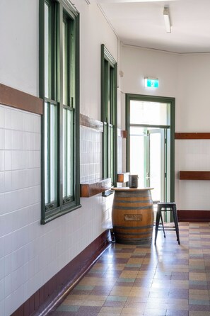 Un pasillo con paredes de azulejos, ventanas con marco verde, un barril de madera y un taburete negro.