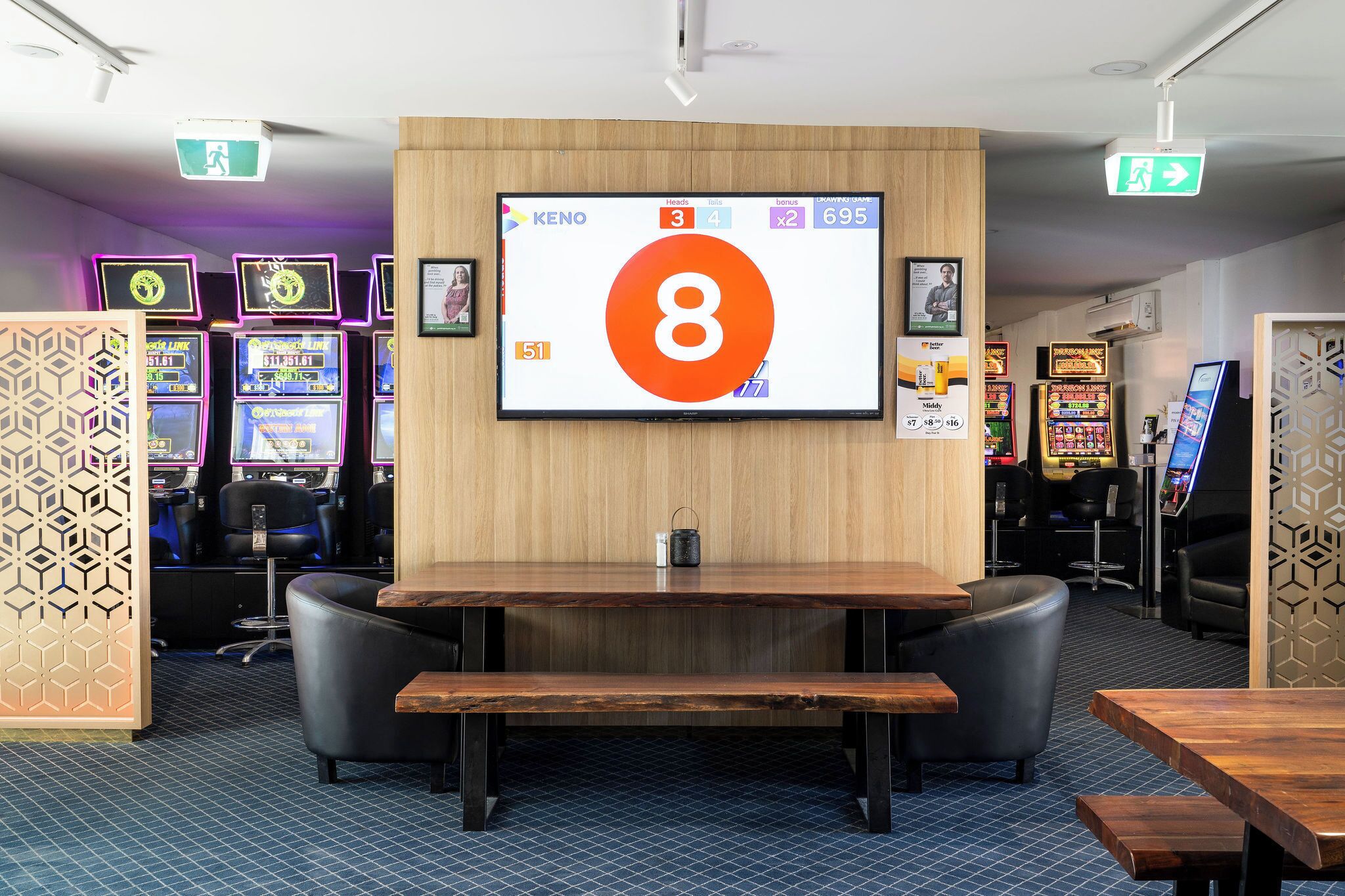 A gaming area with a large screen displaying a game interface, multiple gaming machines, and a wooden table with chairs.