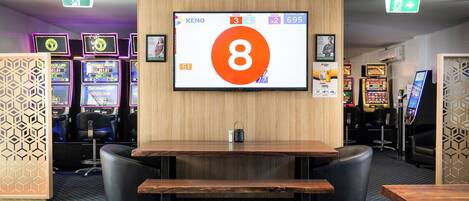 A gaming area with a large screen displaying a game interface, multiple gaming machines, and a wooden table with chairs.