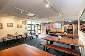 A room with multiple wooden tables and benches, a large screen displaying a game, and framed pictures on the walls. - Queensport Hotel (Brisbane)