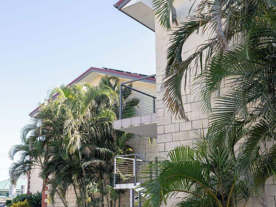 Un edificio con techo rojo, una canasta de baloncesto y abundante vegetación, incluyendo palmeras.