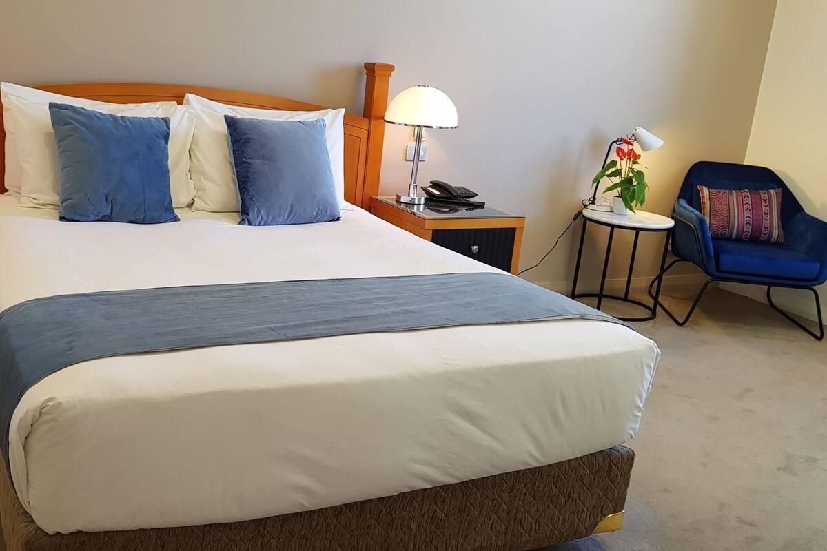 Habitación de hotel con una cama grande, dos mesitas de noche con lámparas, una pequeña mesa con una planta y una silla azul.