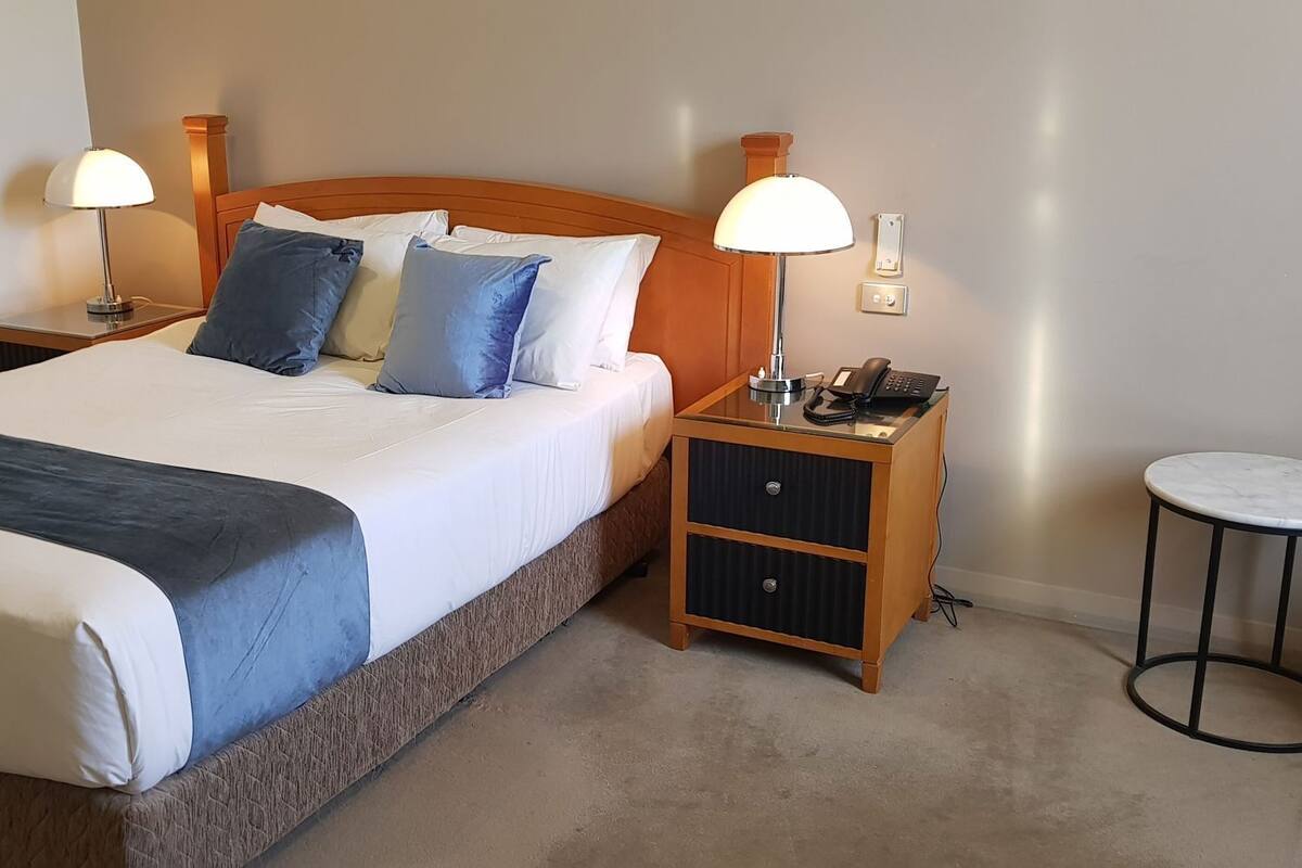 Una cama bien hecha con un detalle azul, dos lámparas de noche, un teléfono junto a la cama, una mesita de noche de madera y una mesita redonda.