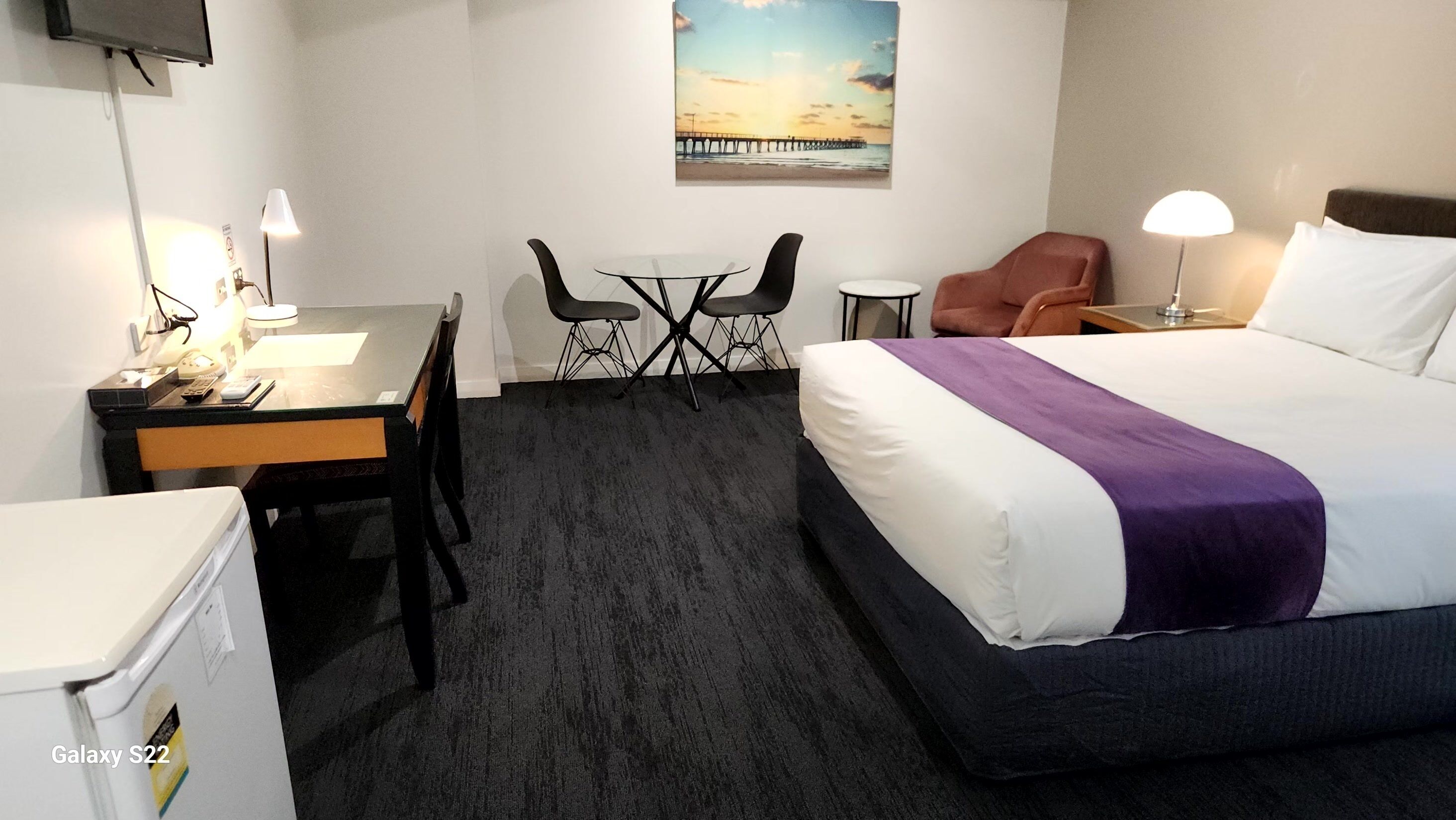 A hotel room with a bed, a desk with a chair, a small table with two chairs, a television, and a painting on the wall.