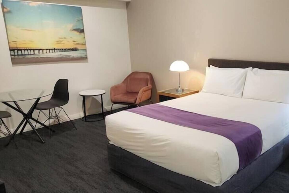 Habitación de hotel con cama, dos lámparas de noche, una silla, una mesita y un cuadro en la pared.