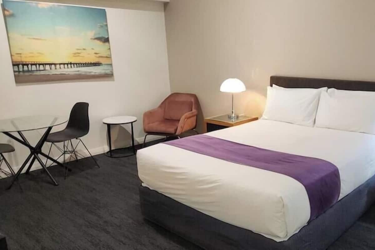 A hotel room with a bed, two bedside lamps, a chair, a small table, and a painting on the wall.