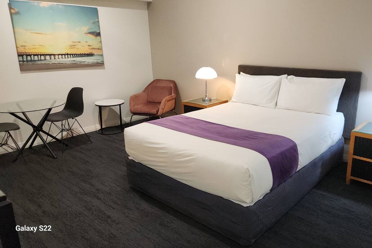 Habitación de hotel con una cama grande, dos mesitas de noche con lámparas, una pequeña mesa redonda con dos sillas, un sillón y un cuadro en la pared.
