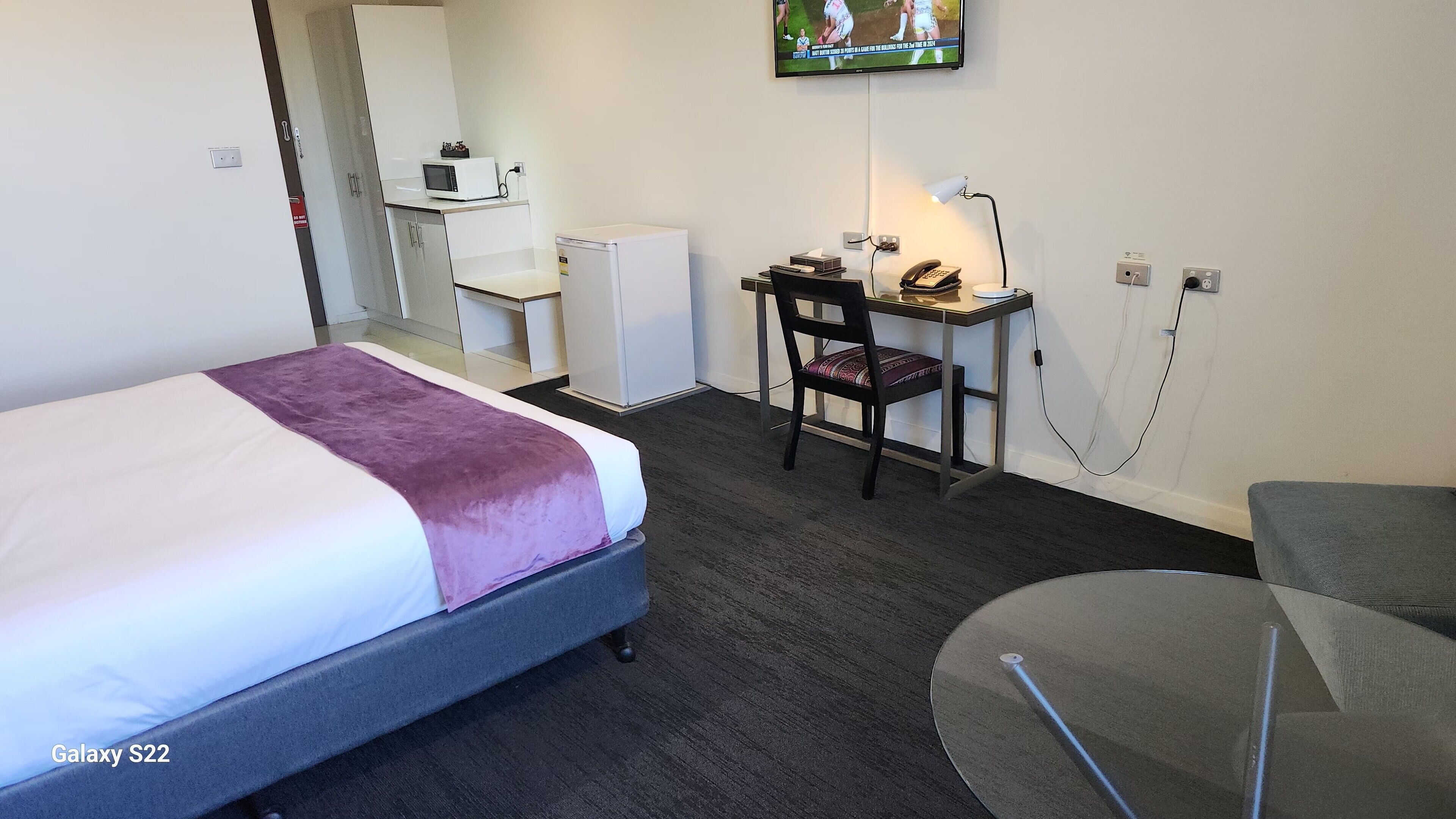 Hotellihuone, jossa on Galaxy S22 -teemalla sisustettu sänky, työpöytä tuolilla, televisio ja pieni pöytä.