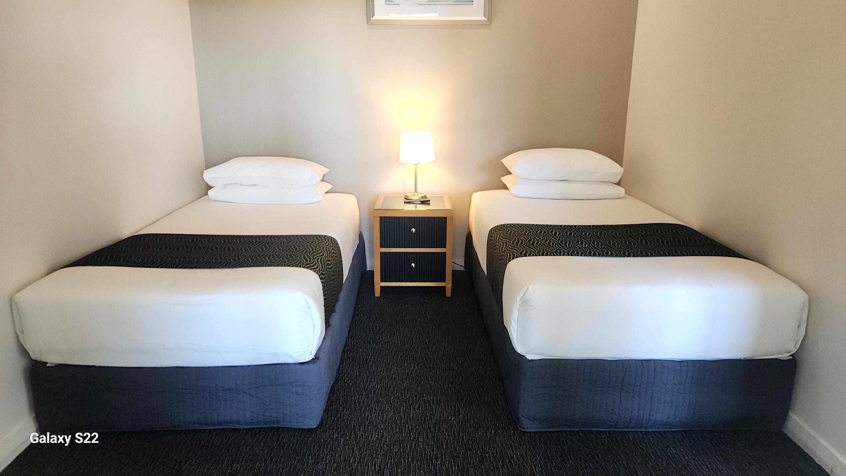 Una habitación de hotel con dos camas individuales, una mesita de noche con lámpara y piso alfombrado.