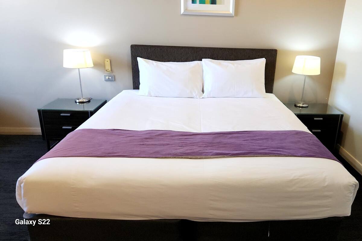Una cama bien hecha con ropa de cama blanca y morada, dos mesitas de noche con lámparas y un cuadro en la pared.