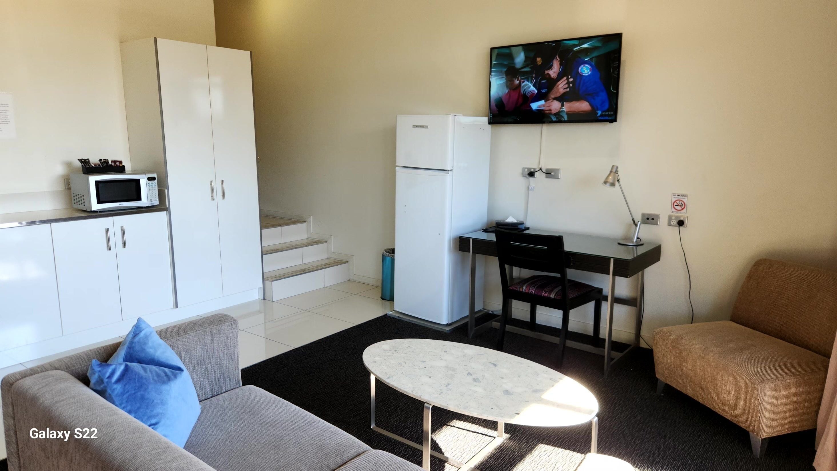 Un espacio habitable compacto con sofá, una mesita, kitchenette con microondas y refrigerador, televisor montado en la pared y una escalera a un nivel superior.
