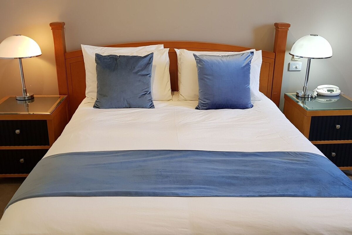 Una cama bien hecha con un detalle azul, dos mesitas de noche con lámparas y un cabecero de madera.