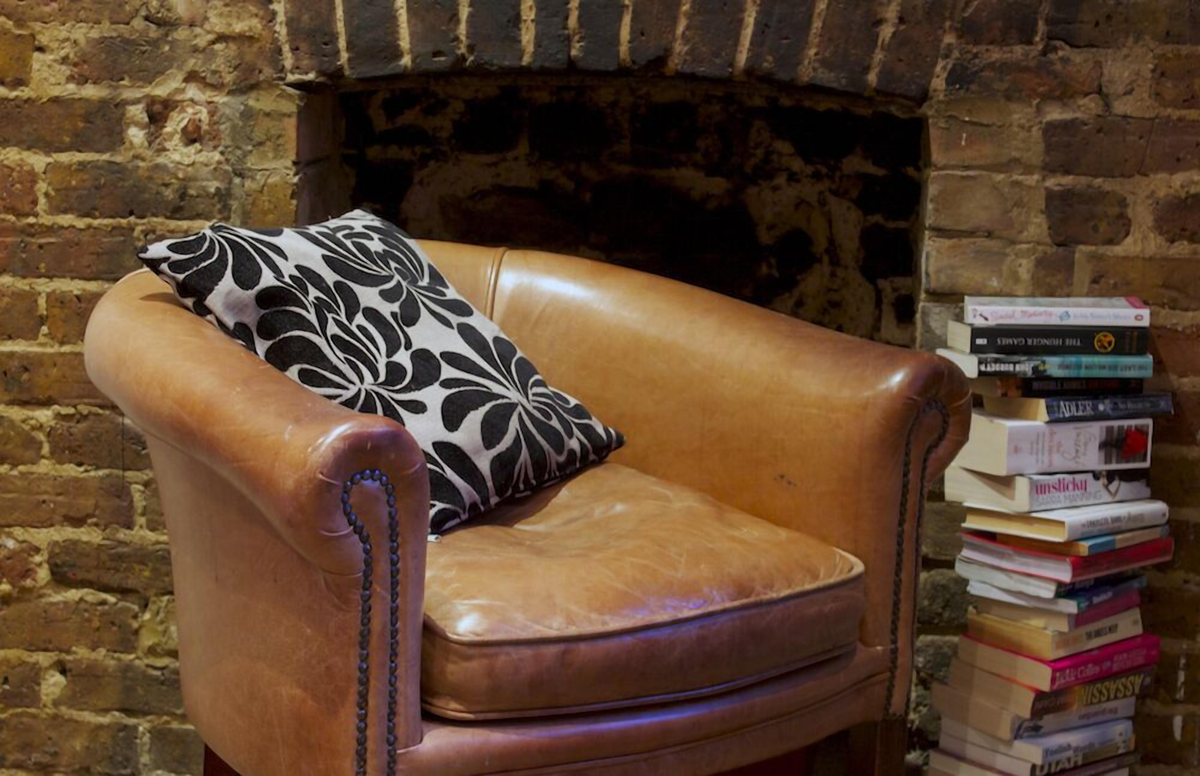 A leather armchair with a black and white patterned pillow, positioned in front of a brick wall with a fireplace. A stack of books is placed to the right of the chair.