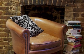 Un sillón de cuero con un cojín estampado en blanco y negro, situado frente a una pared de ladrillo con chimenea. Una pila de libros se encuentra a la derecha de la silla.