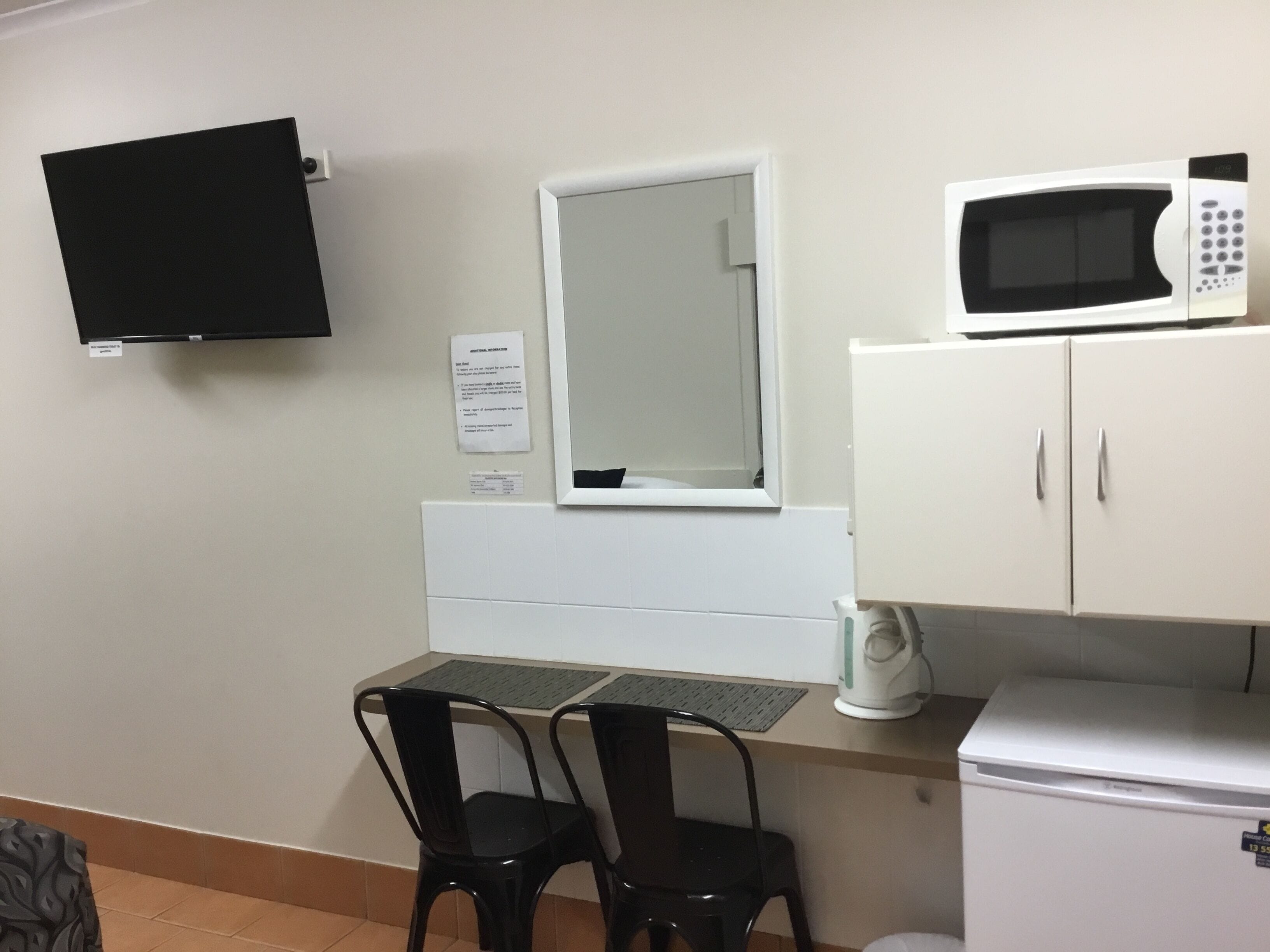 A small kitchenette with a microwave, a refrigerator, a sink, and a window with a mirror. There are two chairs and a table with a keyboard.