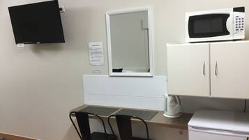A small kitchenette with a microwave, a refrigerator, a sink, and a window with a mirror. There are two chairs and a table with a keyboard.