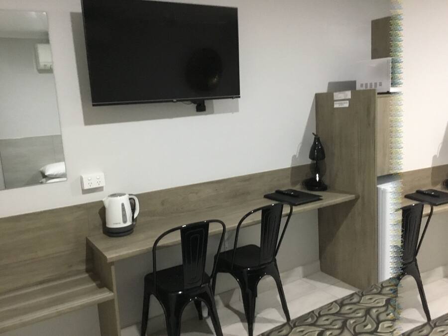 A hotel room with a flat-screen TV mounted on the wall, a wooden counter with a kettle, two black chairs, and a patterned rug.