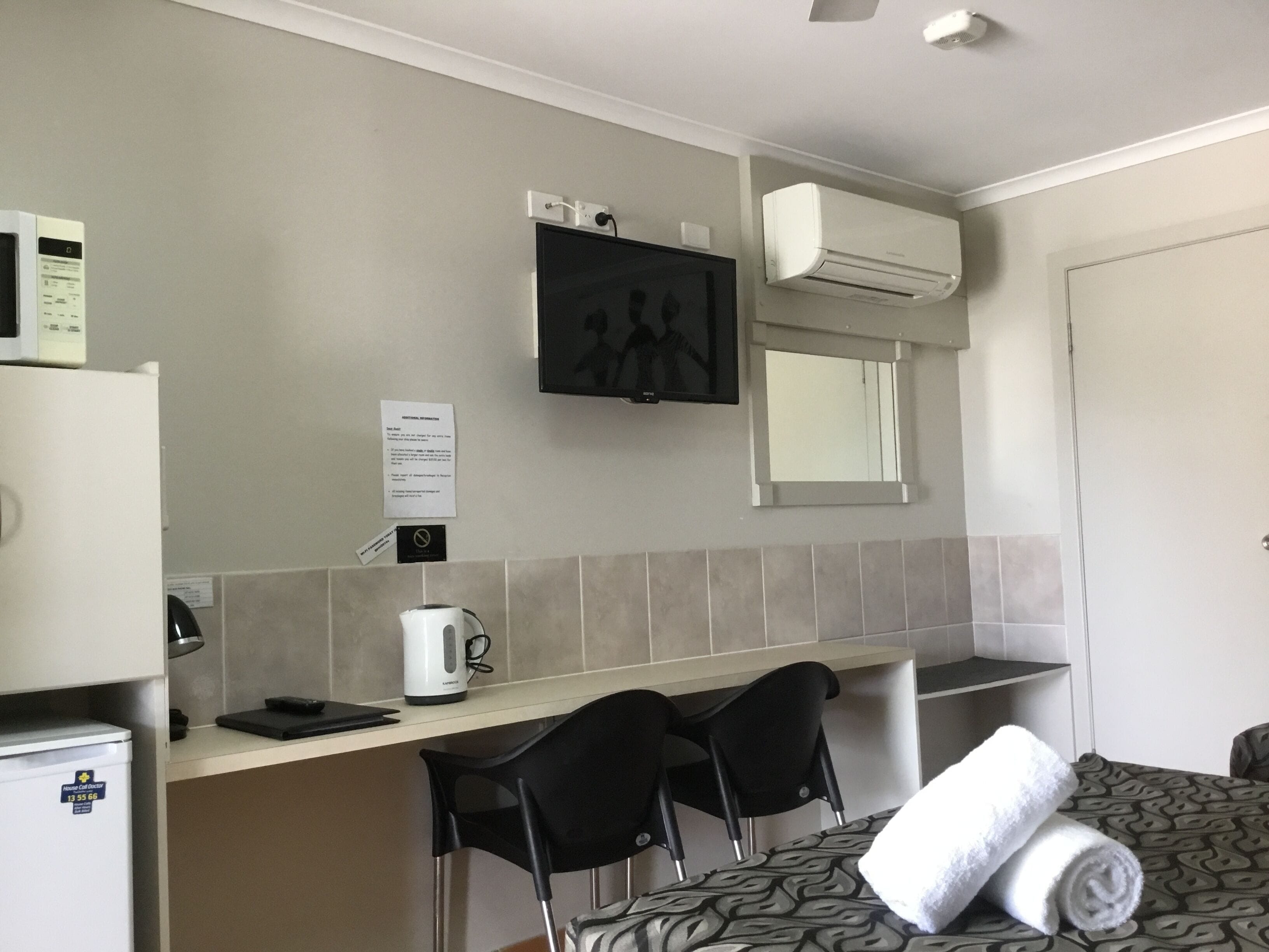 A small kitchenette with a refrigerator, a coffee maker, and a microwave. There is a small table with two chairs and a bed with a rolled-up towel on it.