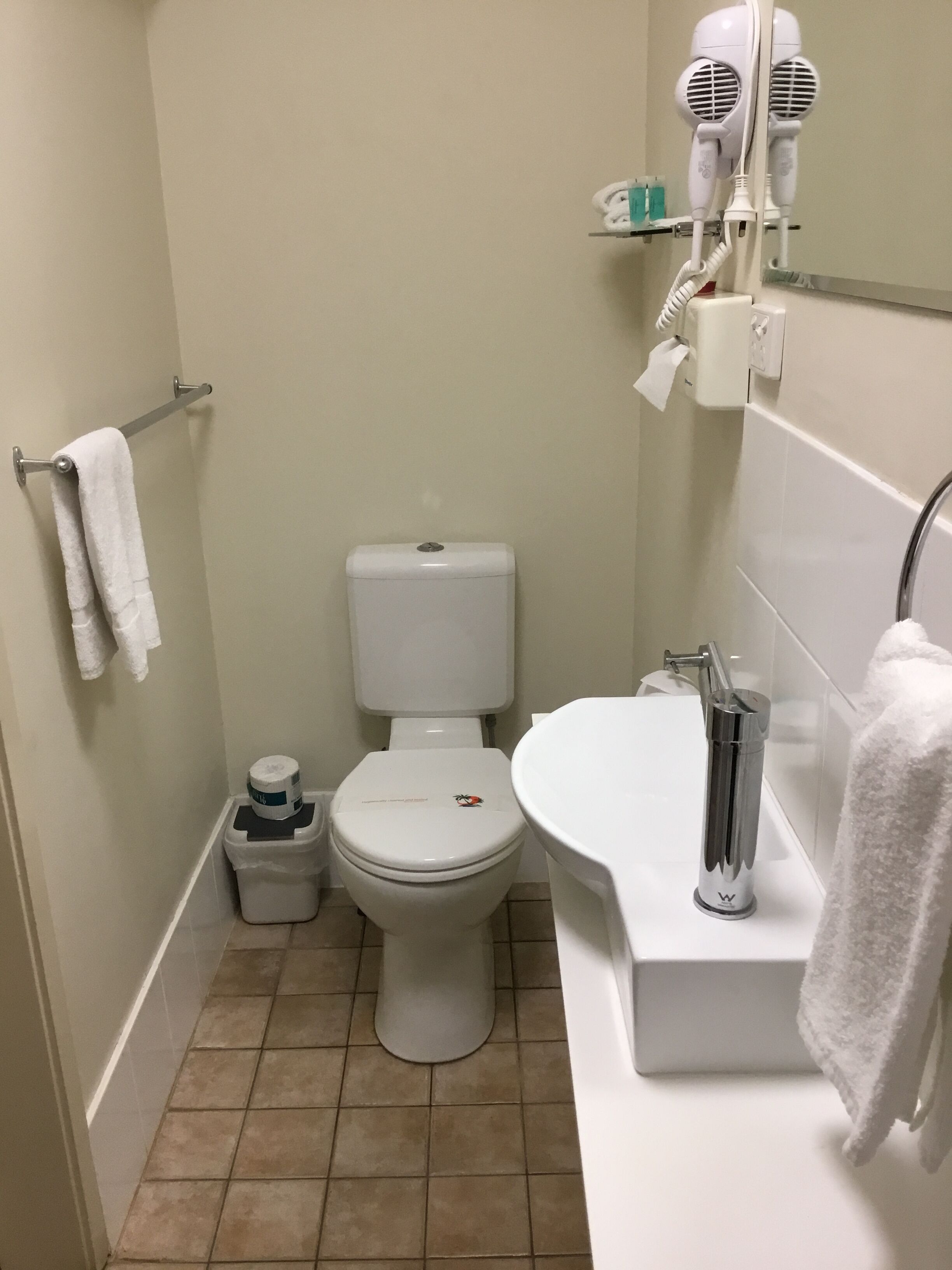 Une petite salle de bain comprenant des toilettes, un lavabo, un porte-serviettes et un sèche-cheveux posé sur une étagère.