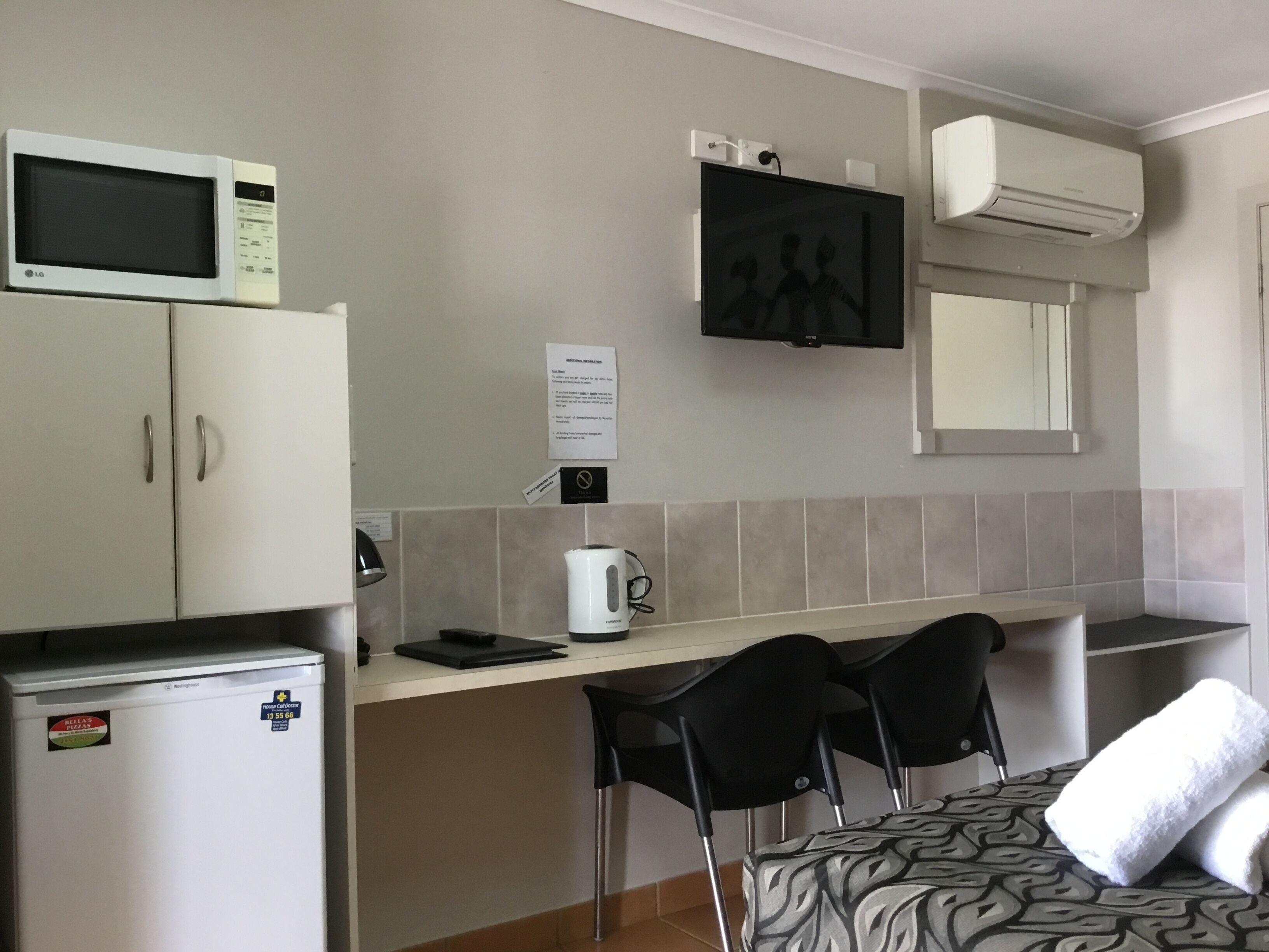 A compact kitchenette with a microwave, refrigerator, and a small coffee maker. There is a flat-screen TV mounted on the wall and a white air conditioning unit above it.