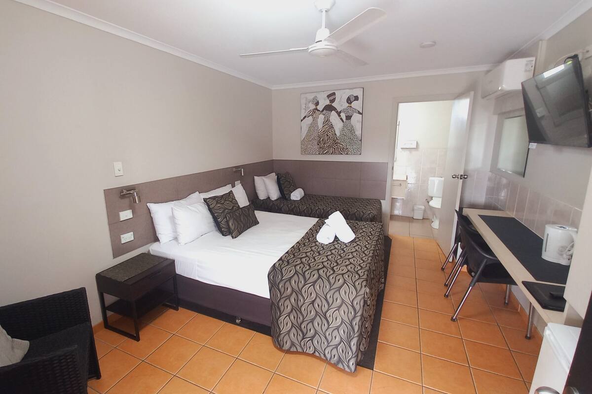 Quarto de hotel com cama, mesas de cabeceira, escrivaninha, cadeira, televisão e banheiro.