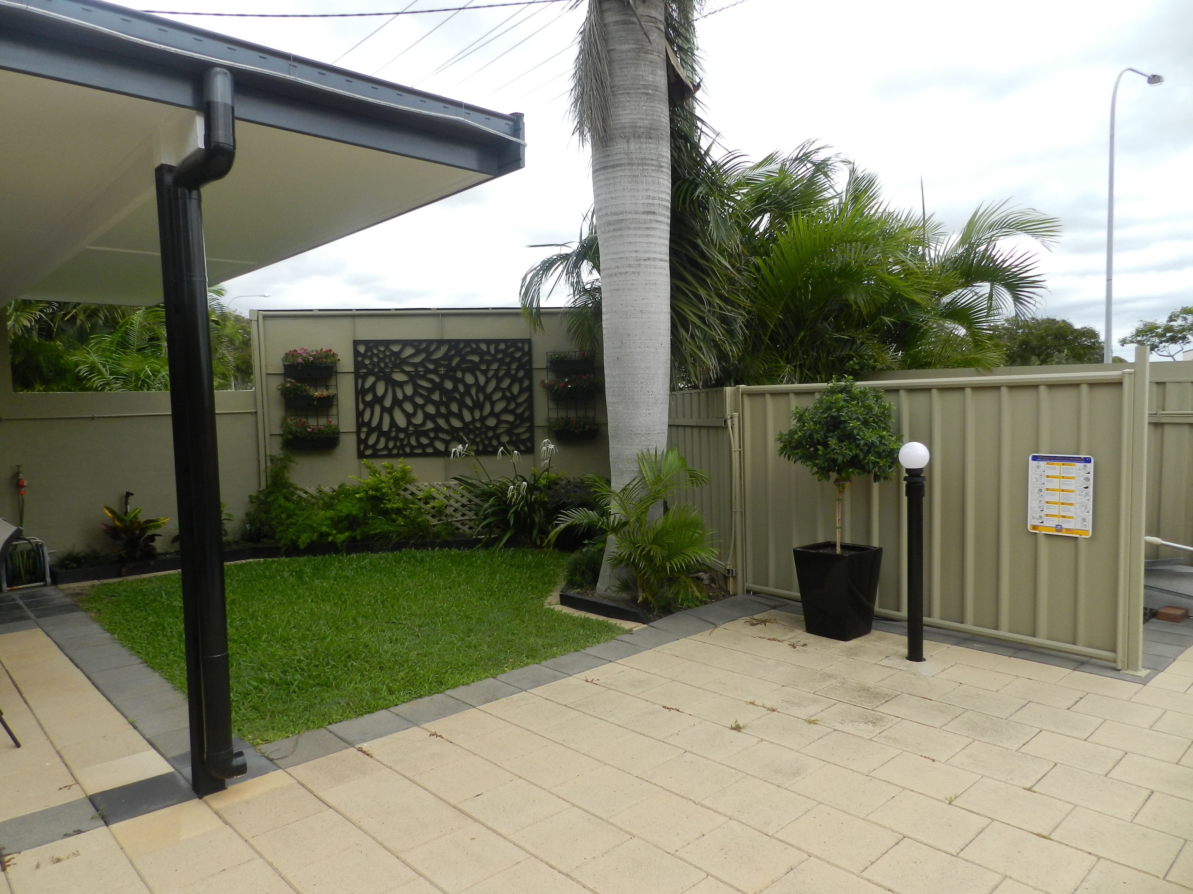 A covered outdoor area with a tiled floor, a palm tree, and a fence with greenery.