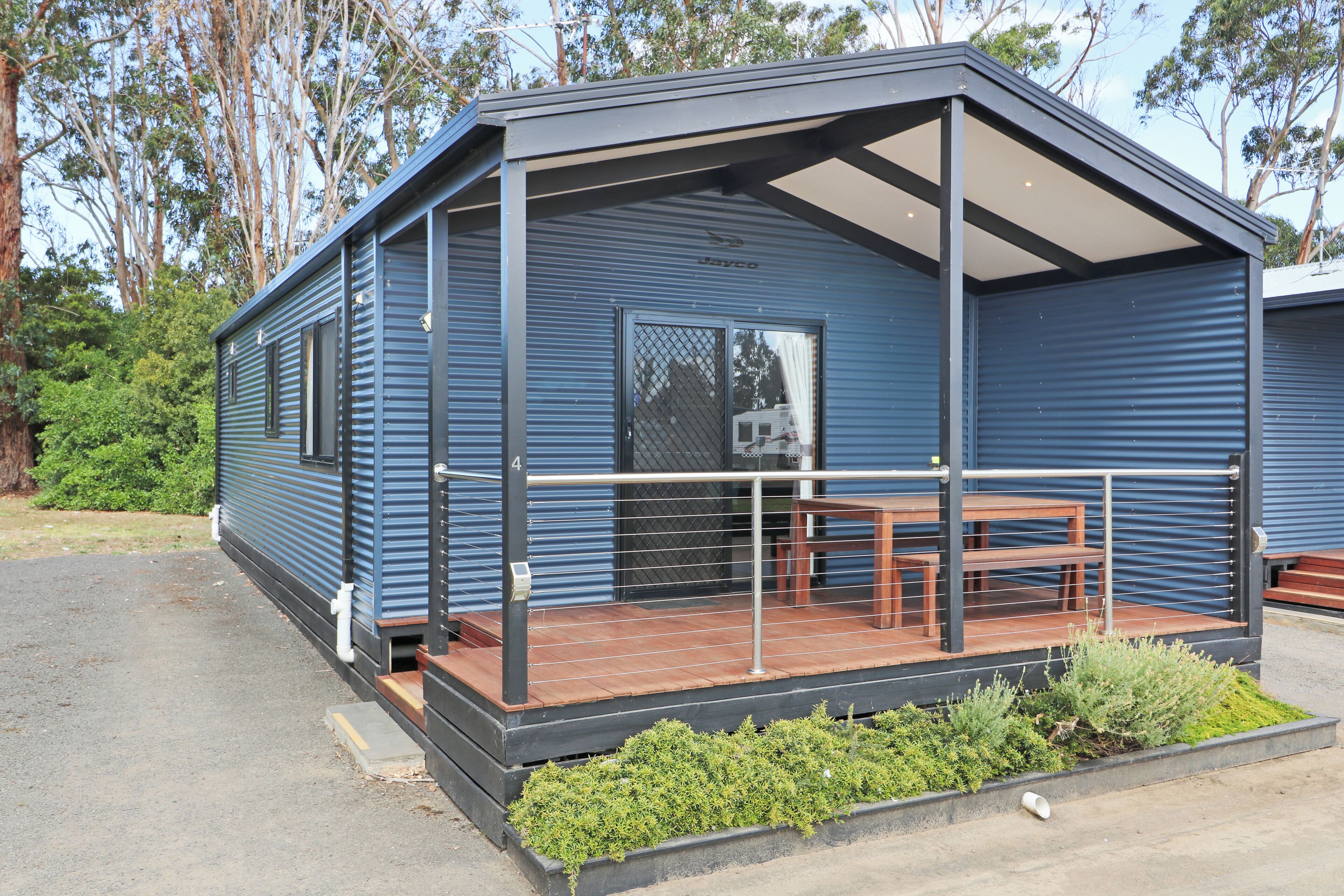 A modern blue mobile home with a wooden deck and a covered outdoor seating area.