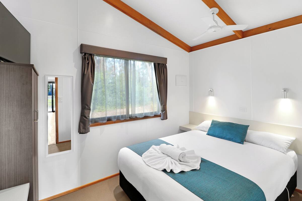 Une chambre d’hôtel comprenant un lit, une table de chevet, une fenêtre avec des rideaux, un ventilateur de plafond et une poutre en bois au plafond.