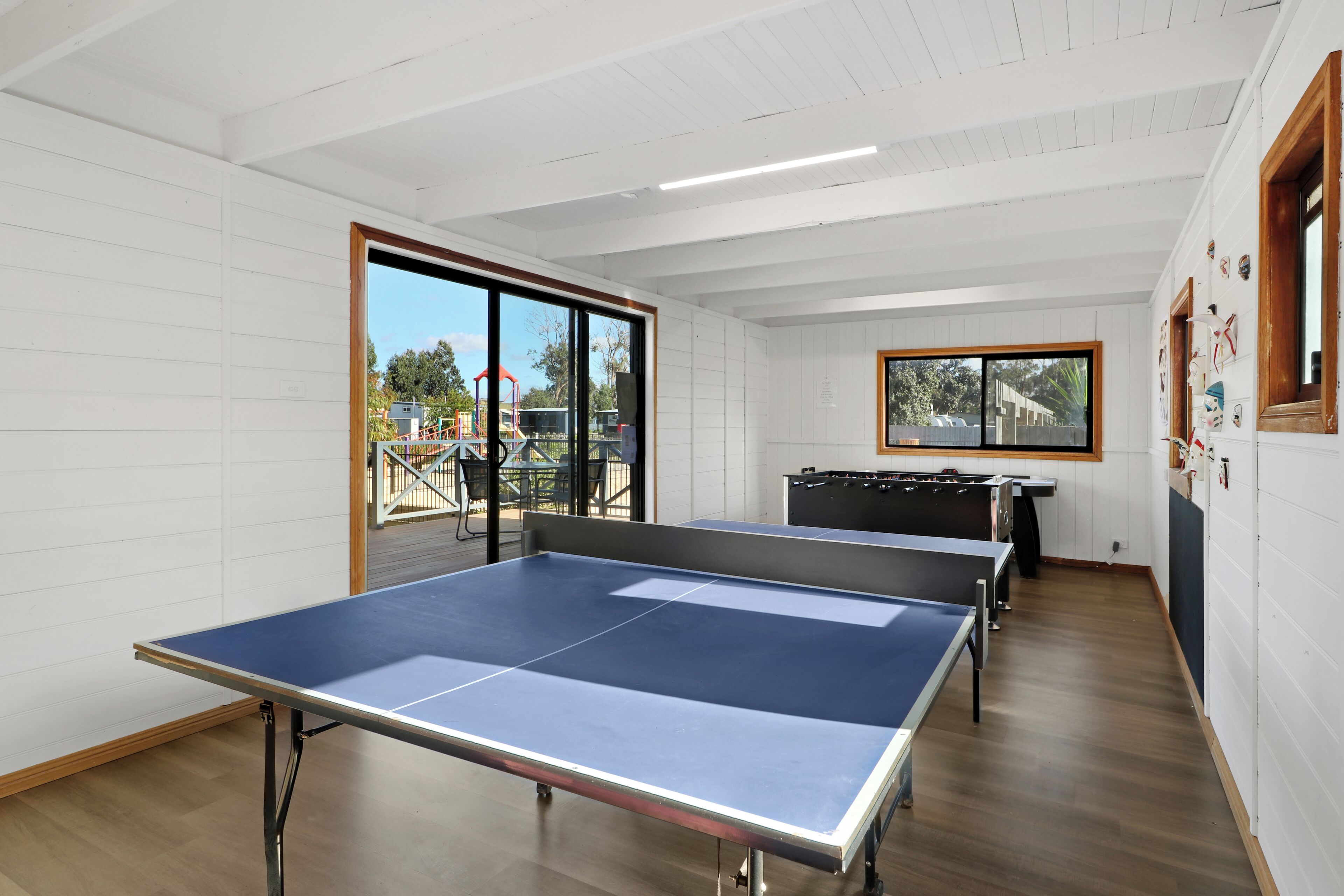 A room with two ping pong tables, a foosball table, and a large window with a view of outdoor play equipment.