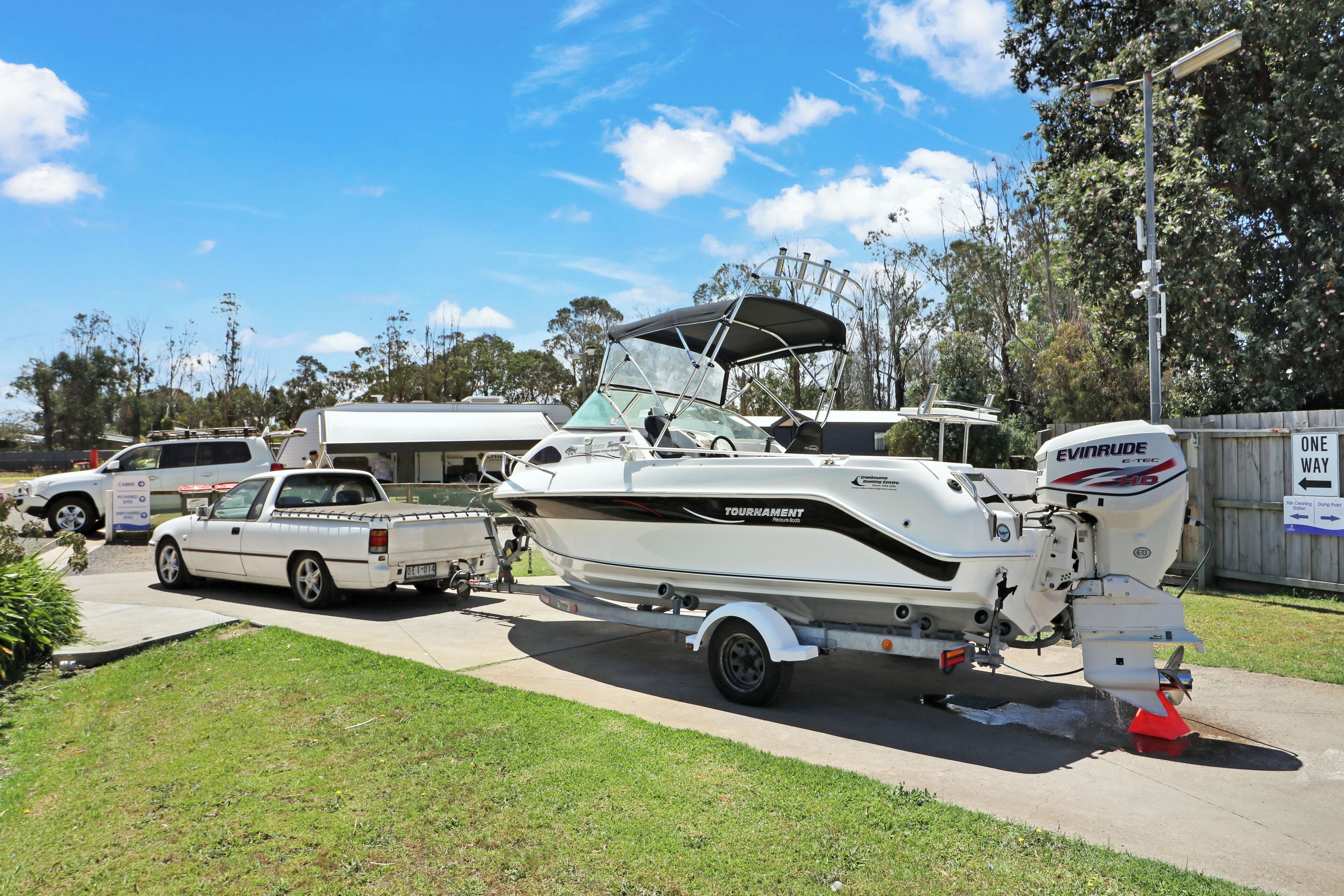 A boat named 'TOURNAMENT' parked on a concrete surface with a trailer, a truck, and a sign indicating 'ONE WAY' in the background.
