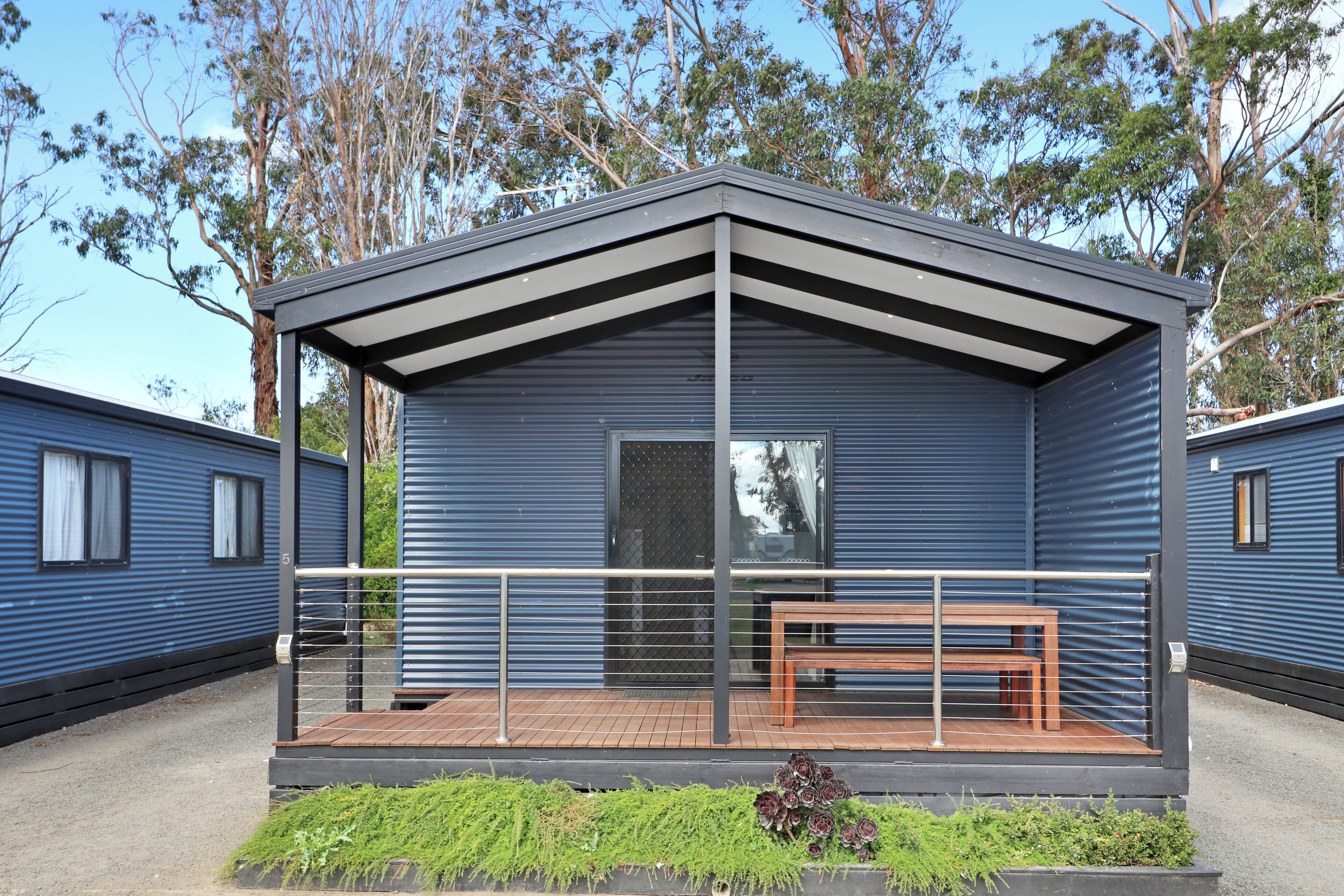 A modern, single-story building with a balcony, a wooden bench, and a small garden area.