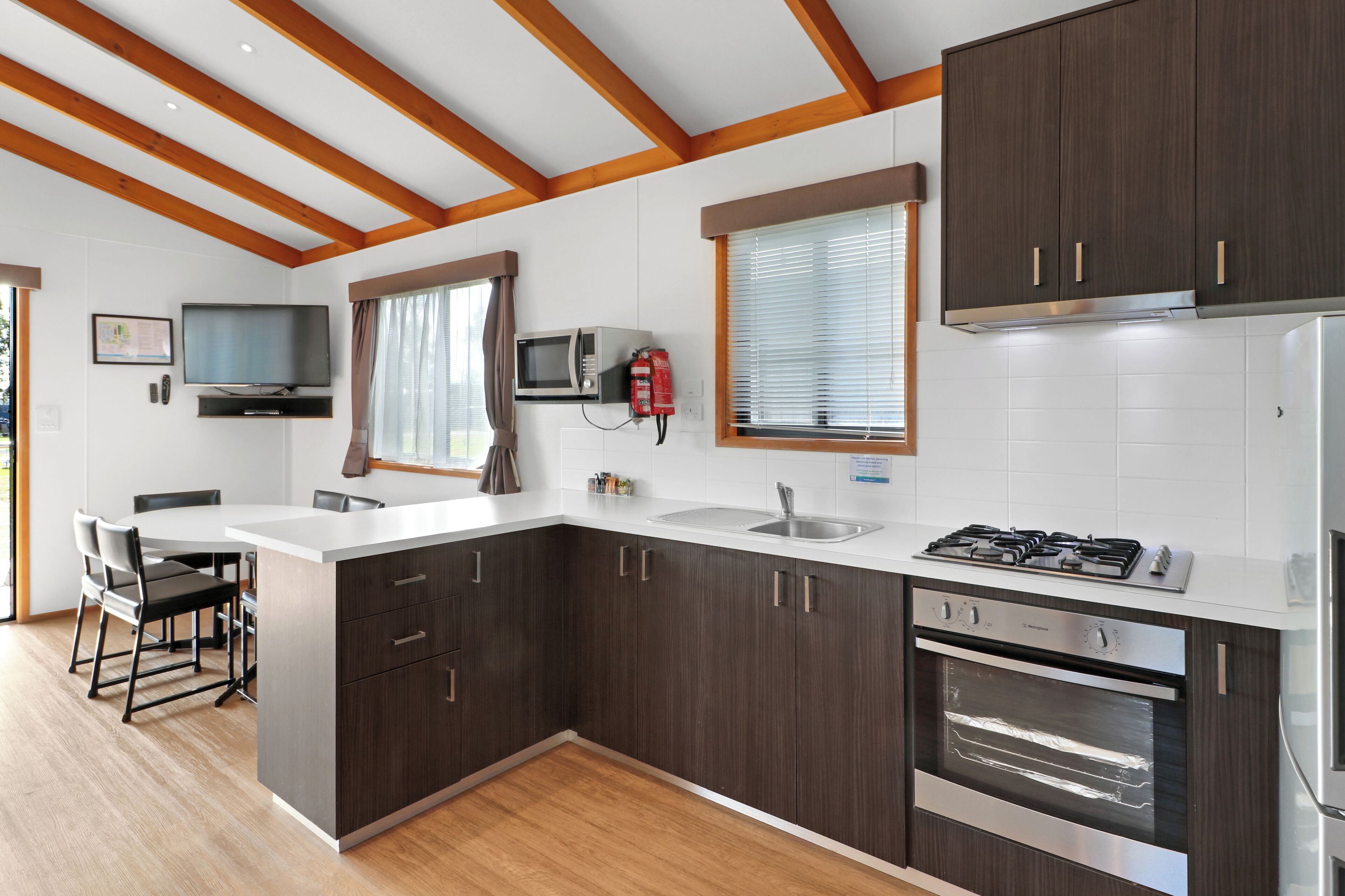 A modern kitchen with dark wood cabinets, a white countertop, a built-in stove, and an oven. There is a dining area with a table and chairs, and a television mounted on the wall.
