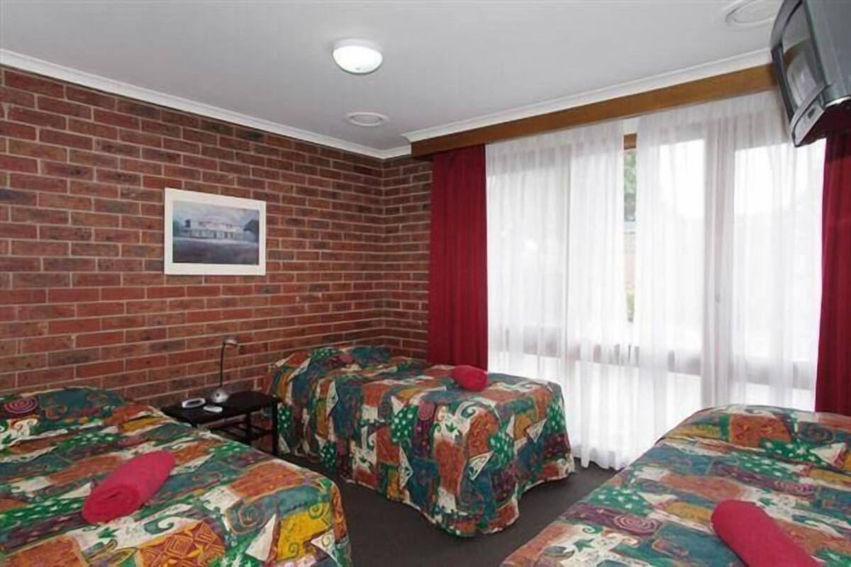A hotel room with two beds, a brick wall, a framed picture, a television, and a window with curtains.