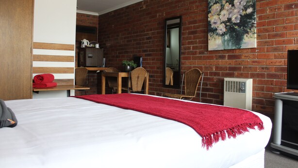 A hotel room with a large bed, a red throw blanket, a brick wall, a painting, a television, and a desk with a chair. - Kardinia Park Motel (Newtown)