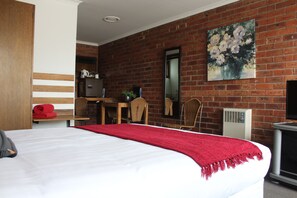 A hotel room with a large bed, a red throw blanket, a brick wall, a painting, a television, and a desk with a chair. - Kardinia Park Motel (Newtown)