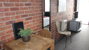 A room with a brick wall, a wooden table with a potted plant, a wicker chair, a bed with a white mattress, a mirror, and a television on a stand. - Kardinia Park Motel (Newtown)