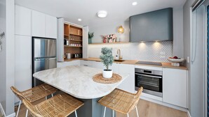 Une cuisine moderne avec des meubles blancs, un réfrigérateur en acier inoxydable, un four encastré et un coin repas avec des chaises en osier.