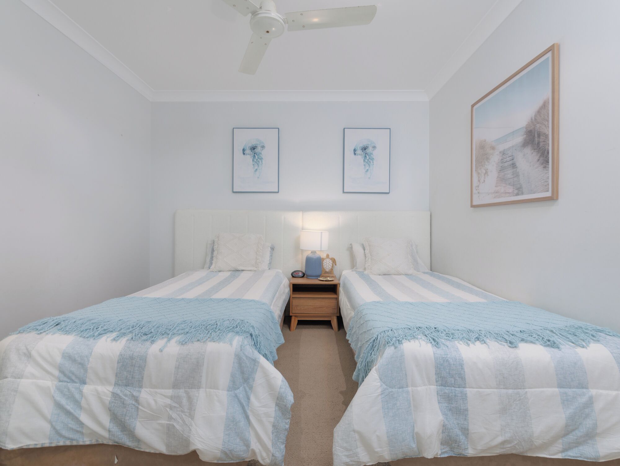 A bedroom with two beds, a ceiling fan, bedside tables with lamps, and framed artwork on the walls.