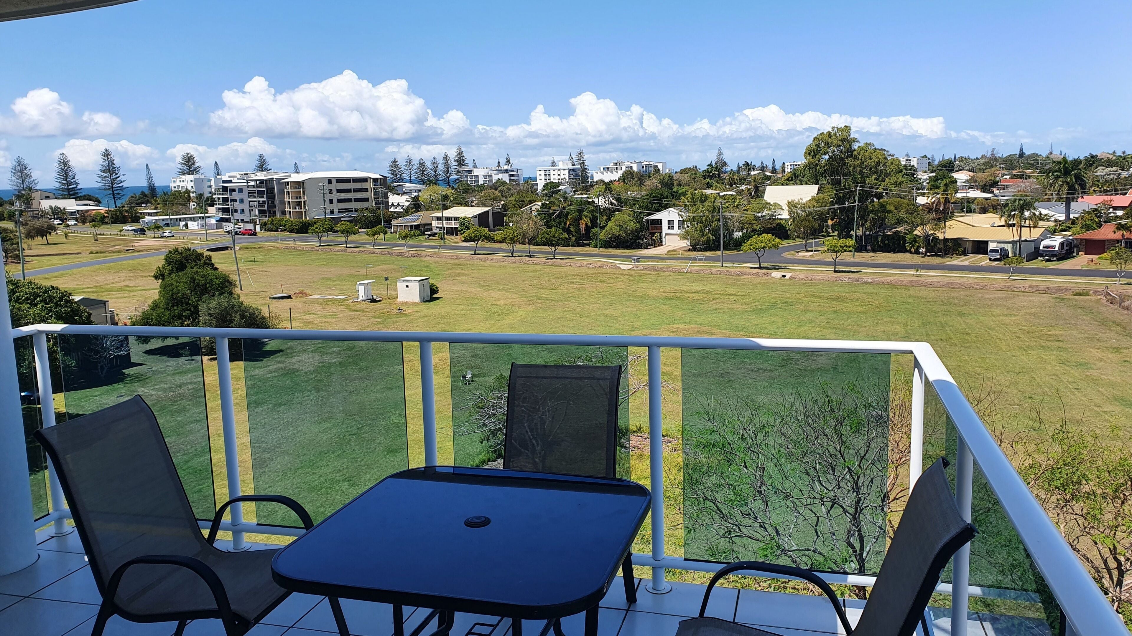 Un balcón con mesa y sillas, con vista a un área verde con edificios y árboles a lo lejos.