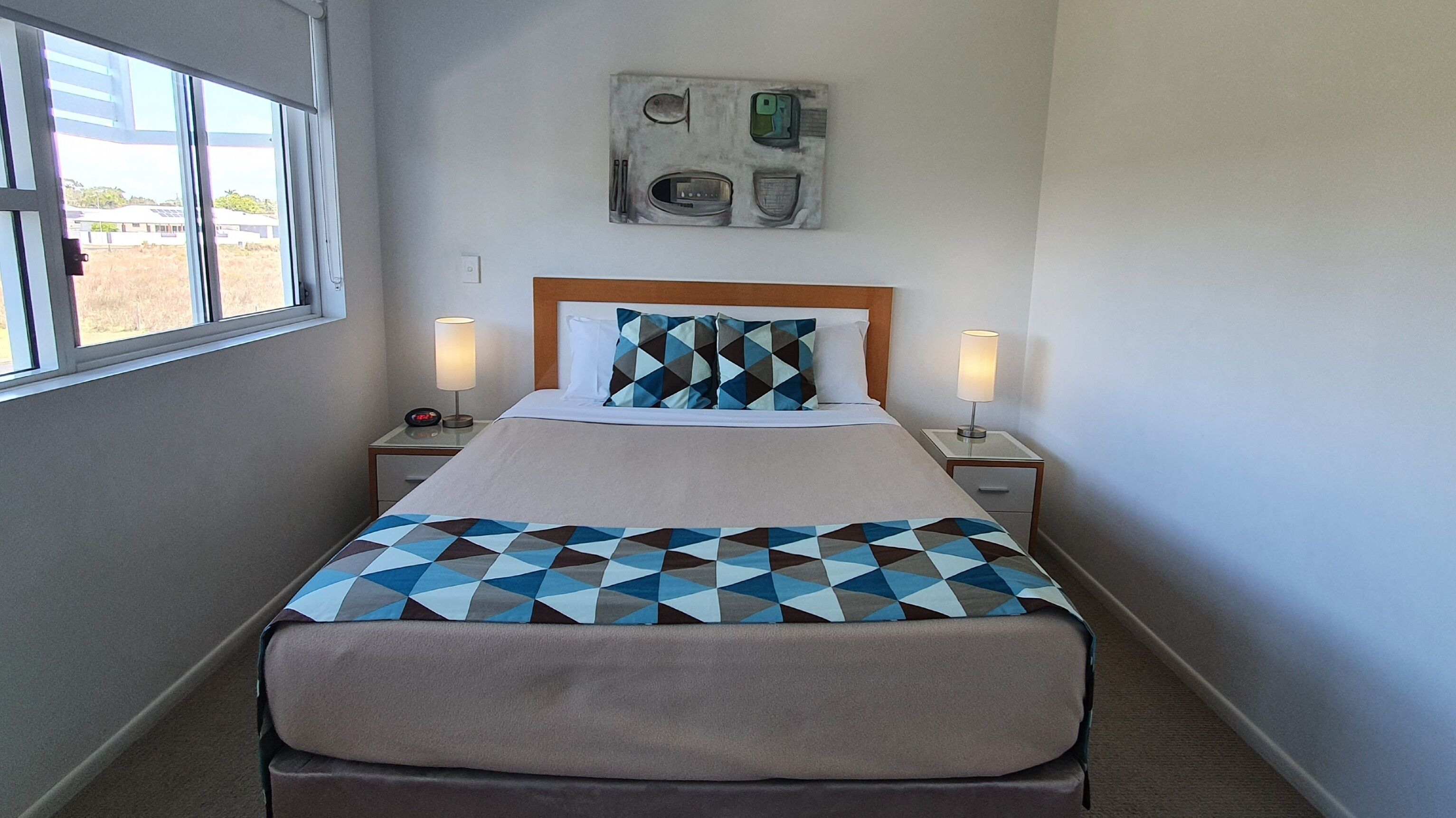 A hotel room with a bed, two bedside tables with lamps, a wall clock, and a geometric-patterned bedspread.