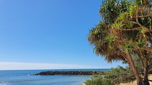 Une plage bordée d’un rivage rocheux, un ciel bleu dégagé et un arbre aux feuilles vertes.