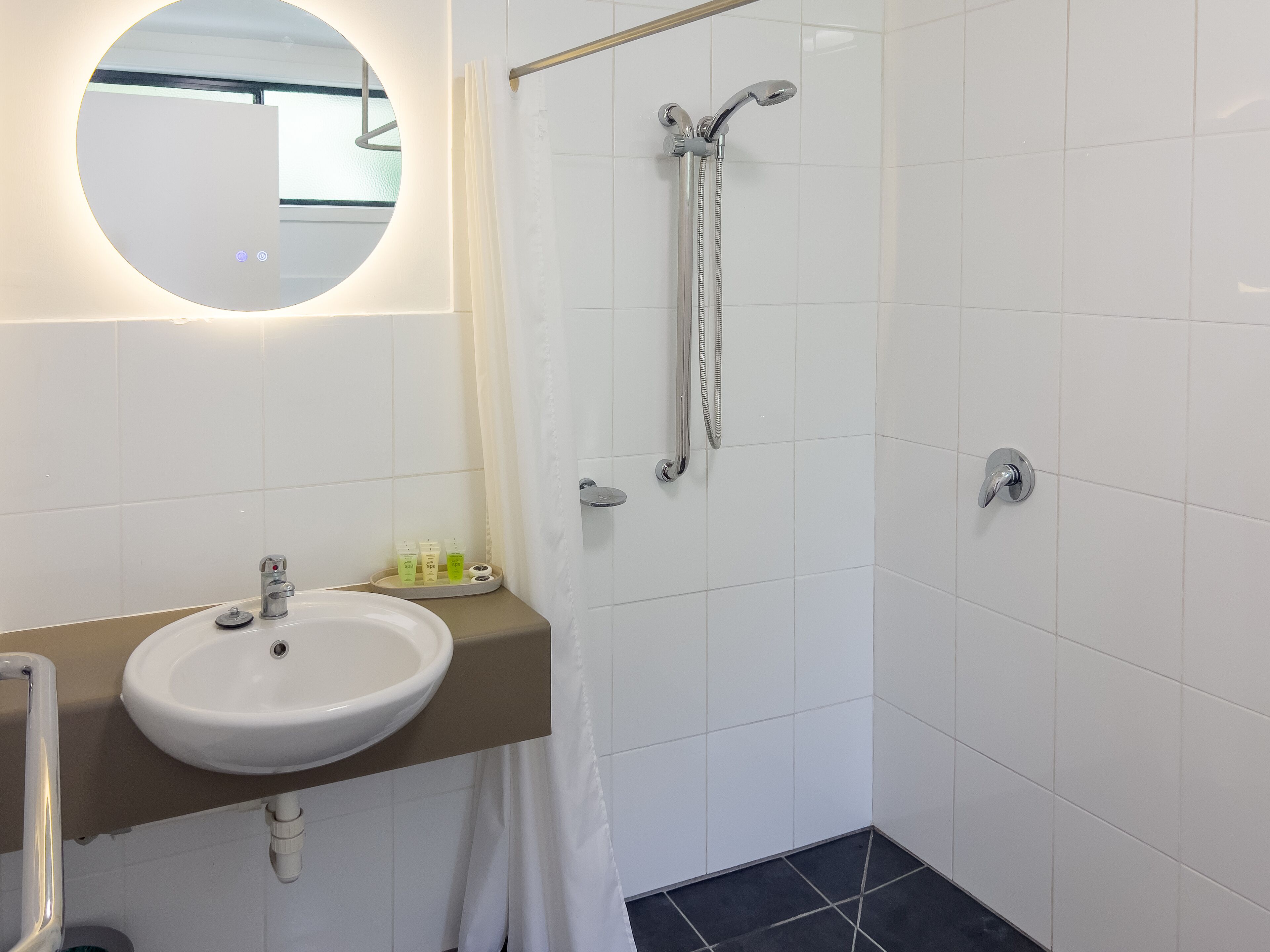 Un baño con lavamanos blanco, espejo, ducha con cabezal de ducha manual y toallero.