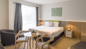 Una habitación de hotel con una cama, una pequeña mesa con sillas, una mesita de noche con lámpara y una ventana con cortinas.