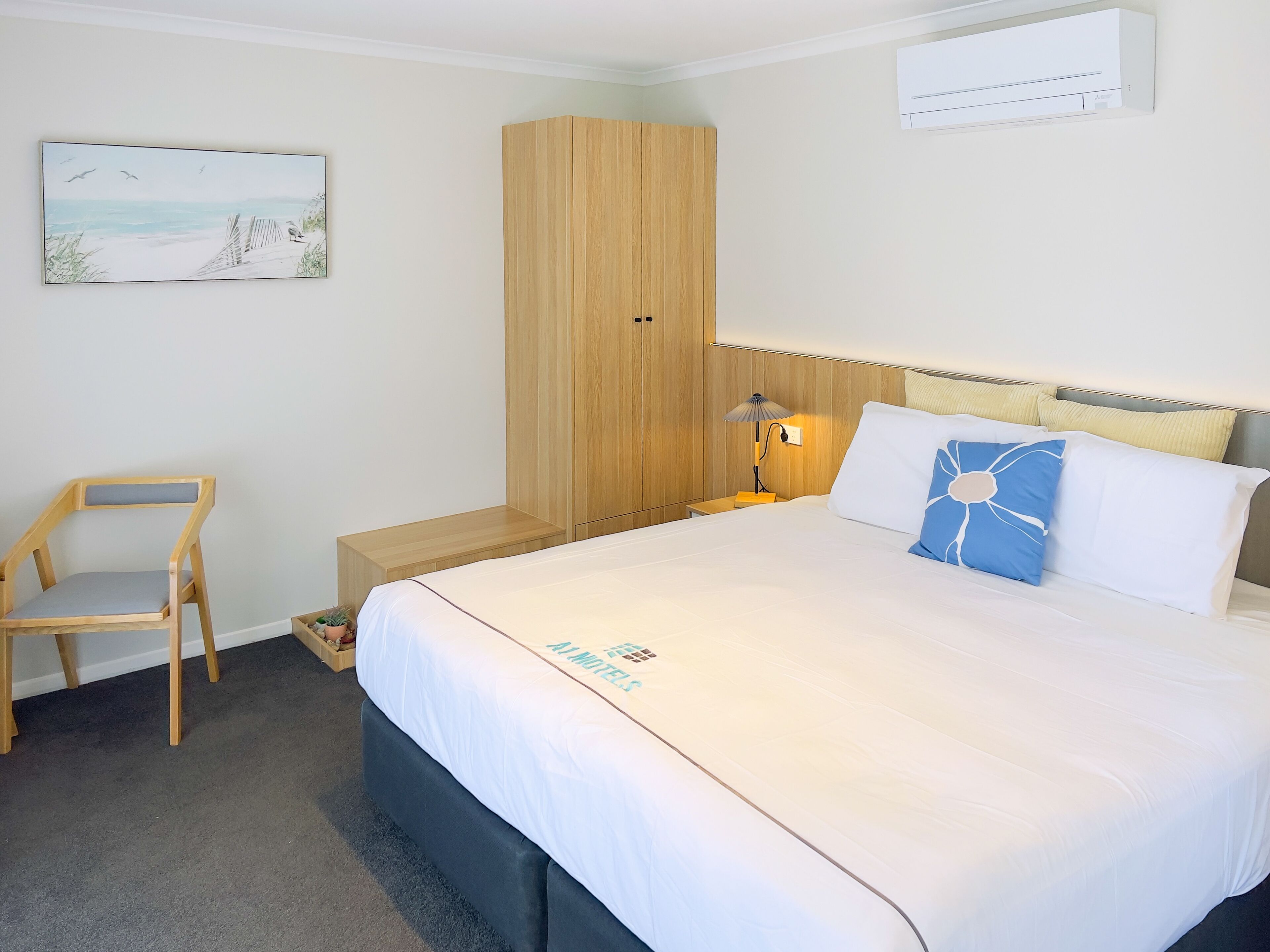 Habitación de hotel ordenada con cama, armario de madera, una silla, mesita de noche con lámpara y aire acondicionado de pared.