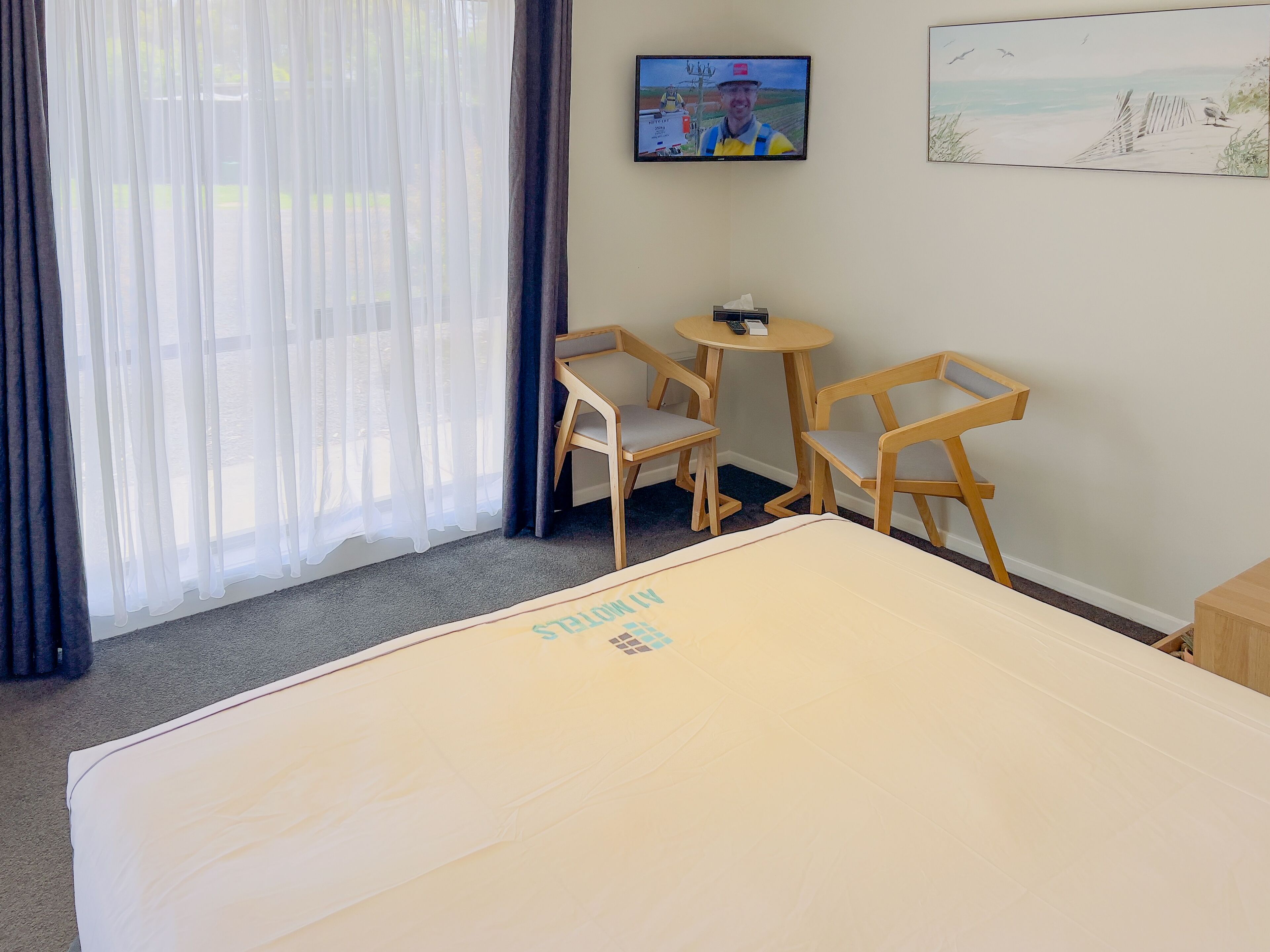 A hotel room with a bed, two wooden chairs, a small table, a TV on the wall, and a painting.
